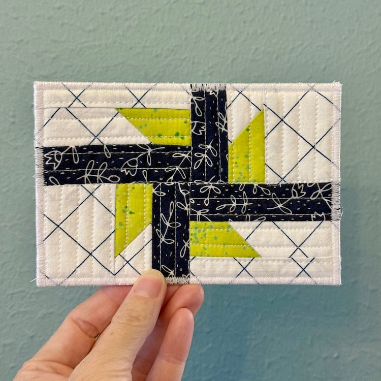 A hand holding a small rectangular quilted postcard in navy, white, and bright yellow-green against a textured light blue wall.