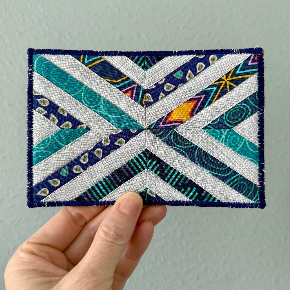 Hand holding a colorful fabric postcard with geometric patterns against a plain background