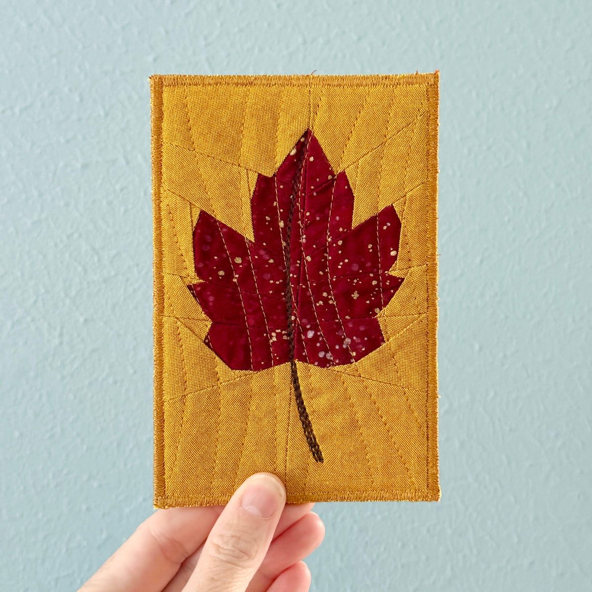 A hand holding a small rectangular quilted block featuring a red maple leaf applique on a mustard yellow background, set against a light blue wall.