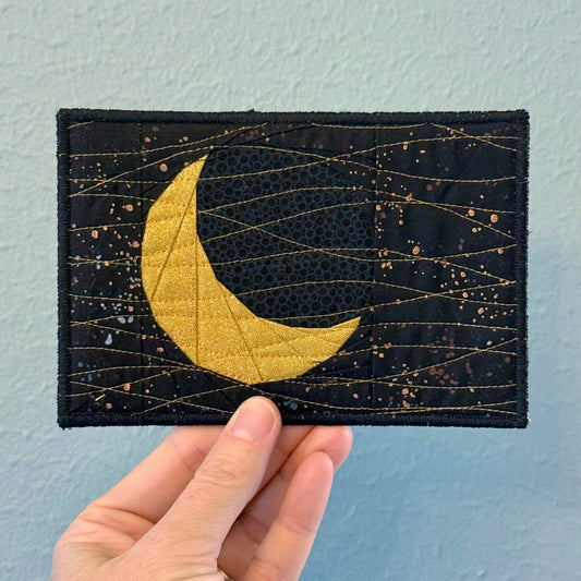 Hand holding a postcard with a gold crescent moon design against a starry black background on a light blue wall.