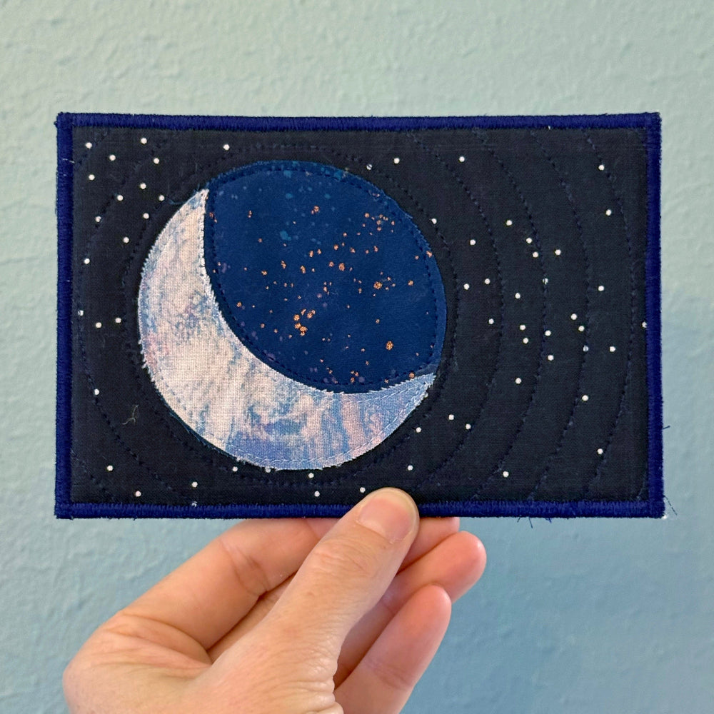 Hand holding a postcard with a blue crescent moon design against a light blue background