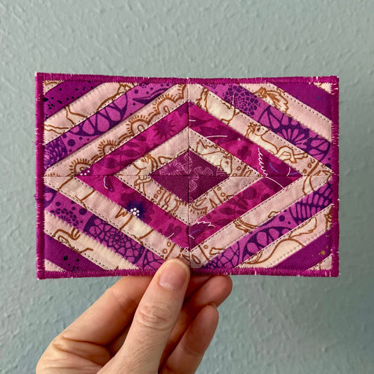 Hand holding a small quilted fabric postcard with purple and pink patterns against a plain background