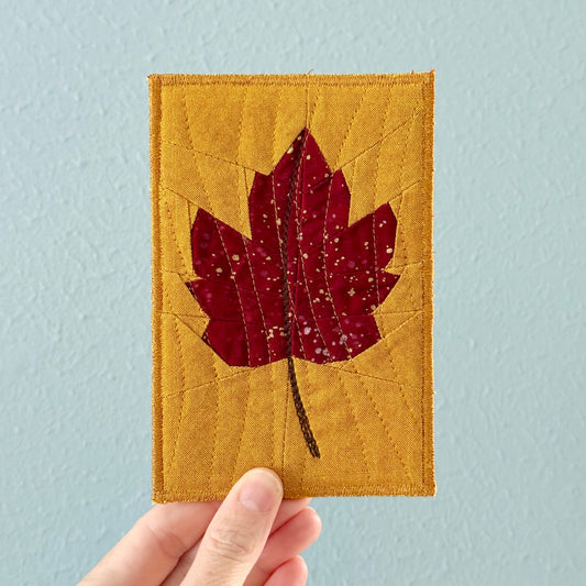 A hand holding a small rectangular quilted block featuring a red maple leaf applique on a mustard yellow background, set against a light blue wall.