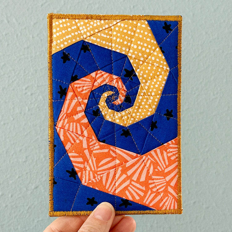 Hand holding a quilted postcard with a spiral design against a gray background