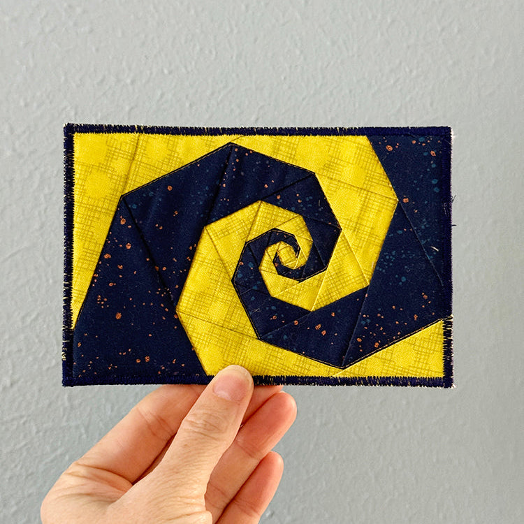 Hand holding a quilted postcard with a yellow and navy blue spiral design against a gray background